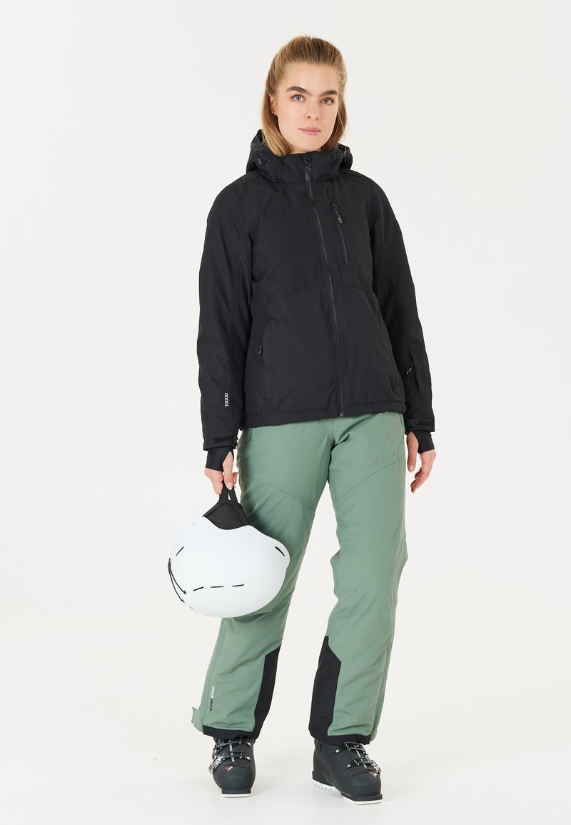 Schwarze wasserdichte Jacke mit Kapuze, kombiniert mit grünen Schneehosen mit schwarzen Akzenten. Die Person hält einen weißen Skihelm in der Hand.