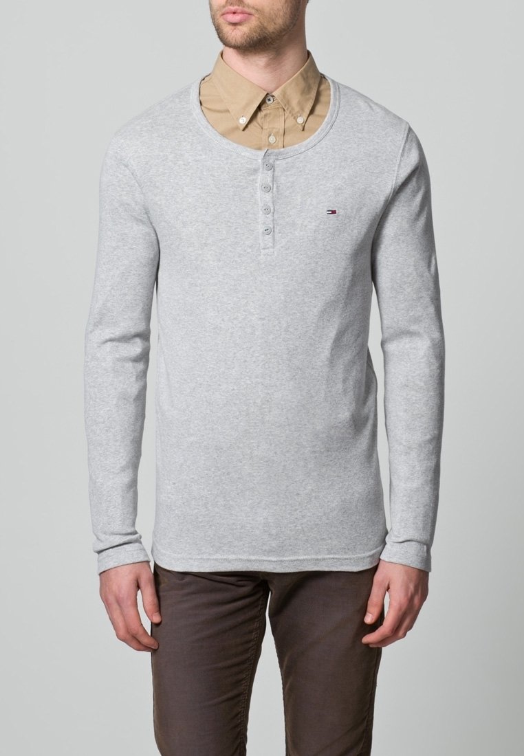 Szara koszulka z długim rękawem, zaokrąglonym dekoltem i trzema guzikami, noszona na beżowej koszuli z kołnierzykiem. Posiada drobny detal z logo.