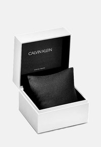 Montre Calvin Klein dans une boîte rectangulaire blanche. L'intérieur est noir avec un coussin noir en velours qui maintient la montre. Texte : "CALVIN KLEIN SWISS MADE."