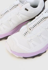 Hvite atletiske sko med nettdesign, svarte og lilla såler, hurtigtrekk-snøring og forsterkede tåhetter.