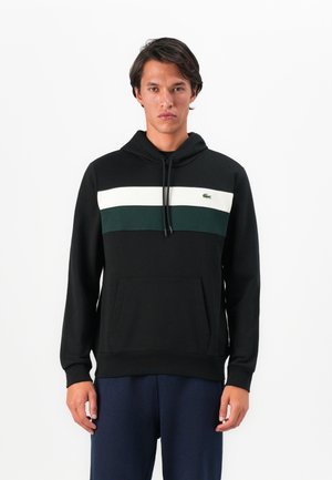 Mann i svart hettegenser med hvite og mørkegrønne horisontale striper og marineblå bukser, stående mot hvit bakgrunn.