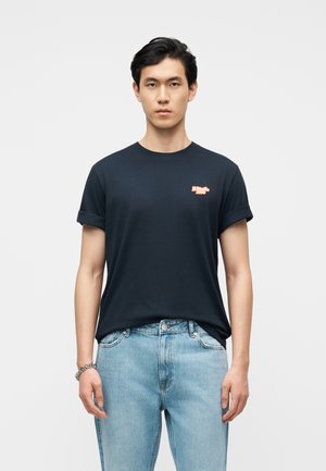 Nuori mies, jolla on mustat lyhyet hiukset, pukeutuneena mustaan T-paitaan, jonka hihat on kääritty ylös, pienellä punaisella logolla, ja vaalean sinisiin farkkuihin, yksinkertaisen taustan edessä.