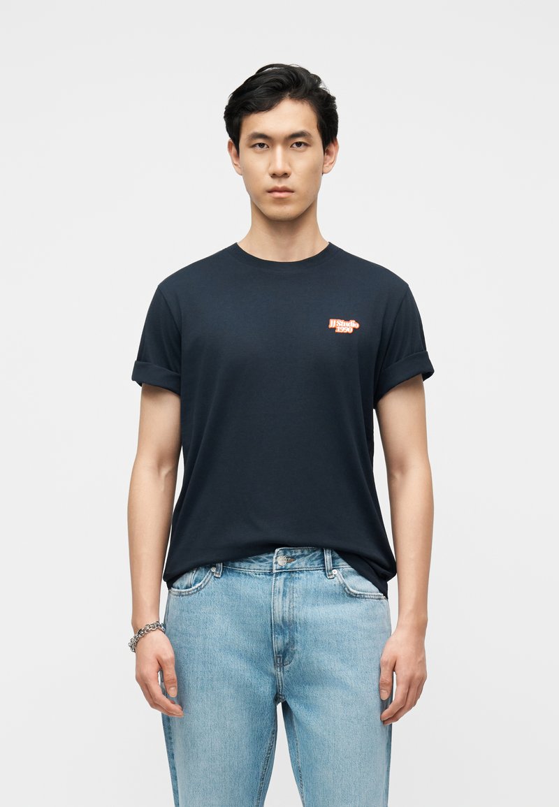 Νεαρός άνδρας με κοντά μαύρα μαλλιά που φοράει μαύρο μπλουζάκι με ανασηκωμένα μανίκια και μικρό κόκκινο λογότυπο, καθώς και ανοιχτόχρωμα μπλε τζιν, μπροστά σε μονόχρωμο φόντο.