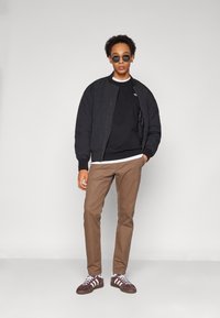 Giubbotto bomber nero sopra una felpa nera, pantaloni marroni e scarpe scure con strisce bianche. Gli accessori includono occhiali da sole rotondi.