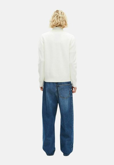 Weißer Strickpullover mit hohem Kragen, kombiniert mit locker sitzenden blauen Jeans. Strukturiertes Material, entspannte Silhouette und Detail mit Gesäßtasche.