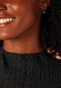 Close-up van een zwart textuur top met een gathered halssnit; heeft een kleine gouden oorring met een heldere edelsteenaccent.