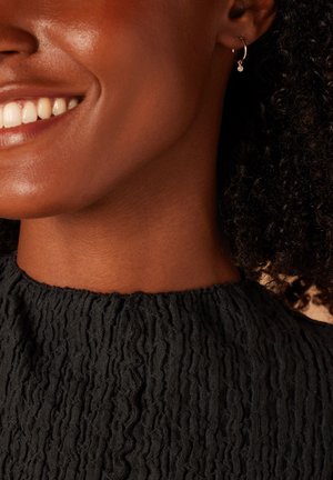 Close-up van een zwart textuur top met een gathered halssnit; heeft een kleine gouden oorring met een heldere edelsteenaccent.