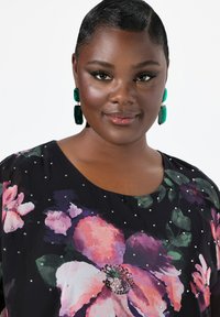 Blusa nera floreale con grandi fiori rosa e viola, decorata con ornamenti metallici. Il modello indossa orecchini geometrici verdi.