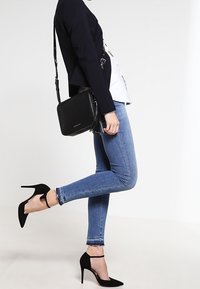 Borsa a tracolla nera con dettaglio di frange, forma quadrata e tracolla regolabile. Indossata con jeans skinny in denim blu e scarpe con tacco nere.