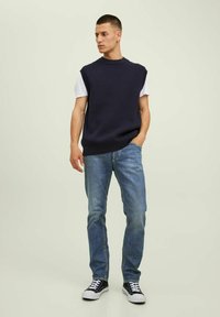 Maglione senza maniche a coste blu navy sopra una t-shirt bianca, abbinato a jeans blu chiaro e sneakers nere, in piedi su uno sfondo neutro.