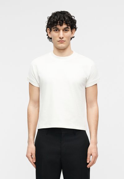 Νεαρός άνδρας με σγουρά σκούρα μαλλιά που φοράει απλό λευκό κοντό μπλουζάκι και μαύρο παντελόνι, στέκεται μπροστά σε ανοιχτό γκρι φόντο.