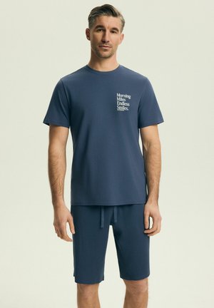 Muž oblečený v tmavomodrom tričku s nápisom "Morning Miles Endless Smiles" a zodpovedajúcich šortkách, stojaci pred jednofarebným svetlým pozadím.