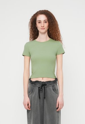 Naine lokkis punaste juustega, kandes helerohelist lühikest t-särki ja lahtisi halli nööriga pükse, seisab tavalise valge tausta ees.