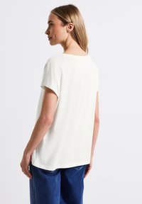 Camiseta blanca holgada de manga corta, con escote redondo y textura suave, combinada con jeans de mezclilla azules. Vista desde atrás.