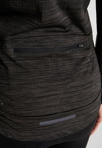 Svart idrottströja med en strukturerad, randig mönstring, med en blixtlåsficka och reflekterande detaljer nära fållen.