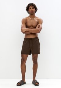 Braune Badeshorts mit elastischem Bund und Kordelzug. Getragen mit dunkelgrauen Slipper-Sandalen. Das Model hat lockige Haare und ist oberkörperfrei.
