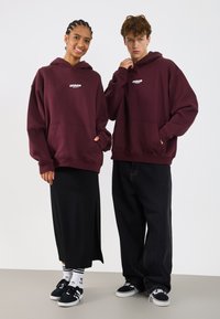 Hanorace bordeaux cu buzunare frontale și logo alb, asociate cu o fustă neagră și blugi lejeri; încălțămintea include adidași negri cu accente albe.