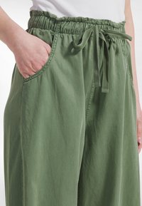 Zelené kalhoty s širokými nohavicemi z měkké látky s elastickým pasem, stahovací šňůrkou a bočními kapsami. Hladká textura a minimalistický design.