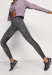 Legginsy Calvin Klein z czarnym i szarym wzorem tekstowym, połączone z dopasowanym fioletowym stanikiem sportowym i jasnofioletową krojoną kurtką.