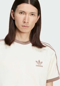 Λευκό βαμβακερό μπλουζάκι με καφέ ριγωτό γιακά και μανίκια. Διαθέτει τρεις καφέ ρίγες στους ώμους και κο embroidered logo της Adidas.