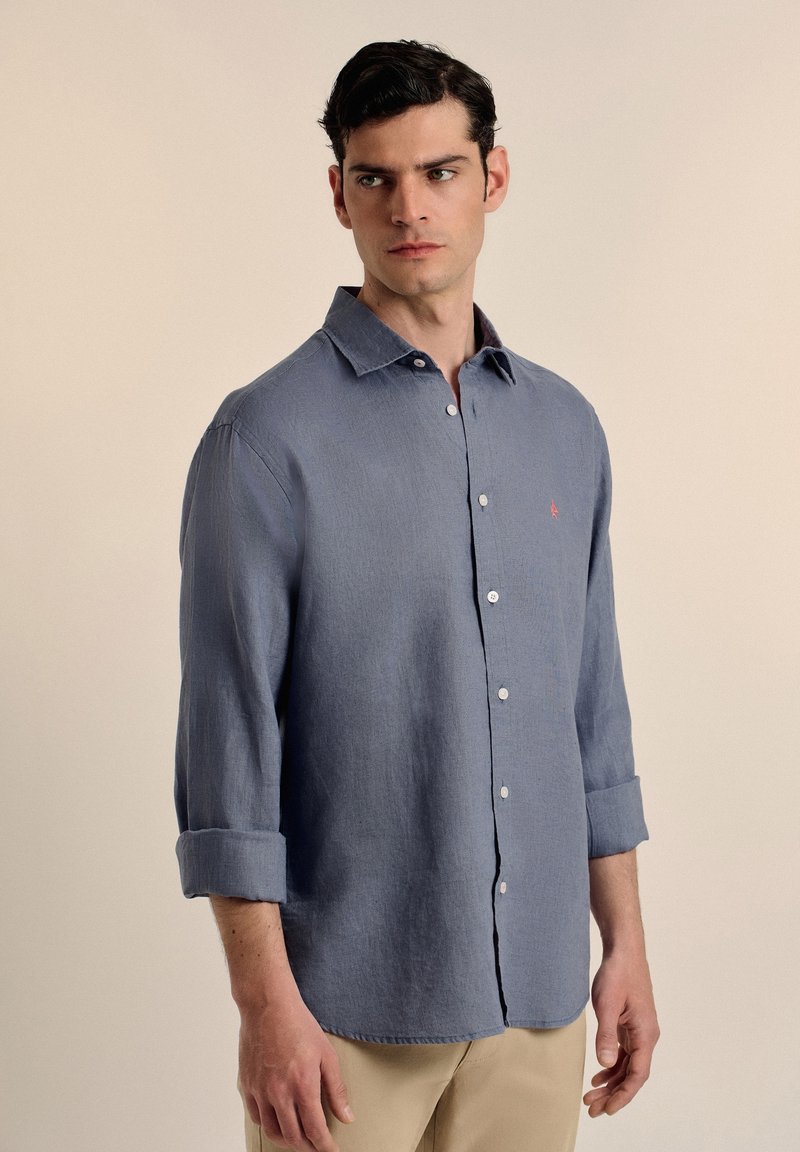 Camisa de botón de manga larga de color azul claro con collar, puños enrollados y un pequeño logo rojo en el pecho. Tejido texturado.