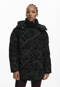 Doudoune noire avec un design matelassé et des motifs de texte audacieux. Col haut, capuche ajustable et poches zippées pour plus de fonctionnalité.