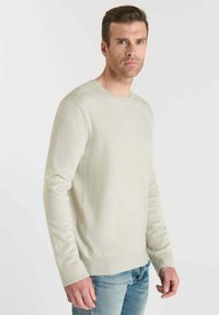 Lichtbeige gebreide pullover met een ronde hals, lange mouwen, ribgebreide manchetten en gestructureerde schouderaccenten, gecombineerd met lichte denim jeans.