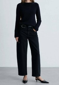 Schwarzer Strickpullover mit rundem Ausschnitt, kombiniert mit weiten schwarzen Jeans mit Gürtel. Dazu werden spitze schwarze Ballerinas getragen.