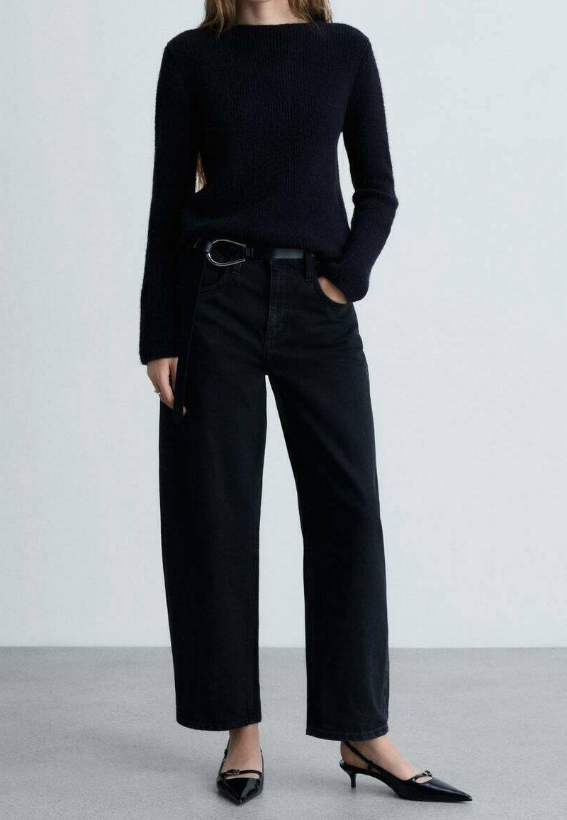 Schwarzer Strickpullover mit rundem Ausschnitt, kombiniert mit weiten schwarzen Jeans mit Gürtel. Dazu werden spitze schwarze Ballerinas getragen.