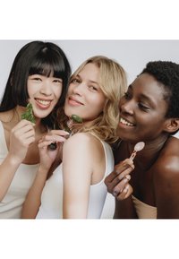 Trois femmes diverses sourient en tenant des outils de massage facial sur un fond neutre.