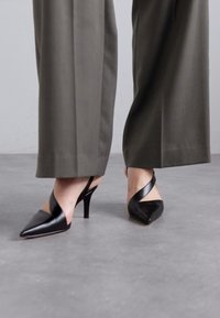 Talons aiguilles noires à bout pointu avec un design élégant, présentant un talon fin et une sangle texturée traversant le pied.
