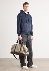 Navy zip-up hoodie with a textured front pocket, gray jeans, and a beige canvas duffel bag with brown leather accents and handles.