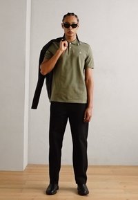 Tricou polo verde măsliniu, pantaloni negri și ochelari de soare negri. Modelul ține o jachetă neagră pe un umăr, stând pe un podea din lemn.