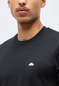 Άνδρας με απλό μαύρο μπλουζάκι λαιμόκοψης με μικρό λευκό ημικυκλικό λογότυπο στο αριστερό στήθος, εμφανιζόμενος από τους ώμους μέχρι τα χείλη.