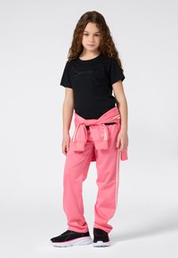 Niña joven con cabello largo y rizado, vistiendo una camiseta negra de "Champion", pantalones de chándal rosas, sudadera rosa atada a la cintura y zapatillas negras.