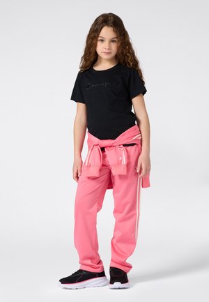 Junges Mädchen mit langen lockigen Haaren, das ein schwarzes "Champion"-T-Shirt, rosa Jogginghose, einen rosa Sweatshirt um die Taille gebunden und schwarze Turnschuhe trägt.