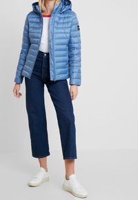 Jasnoniebieska pikowana kurtka z kapturem, z przeszyciami, zapinana na zamek, z naszywką na lewym rękawie. W połączeniu z ciemnymi dżinsami i białymi trampkami.