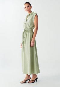 Svetlo zelena brezrokavna maksi obleka z gumbnim zapenjanjem spredaj, z vračeno zapenjalno pasom in elegantnim krojem, v kombinaciji s črnimi klini flip-flopi.