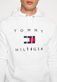 Sudadera blanca con cordones, que presenta un logo con franjas horizontales en rojo, blanco y azul, y el texto "TOMMY HILFIGER" en negro.