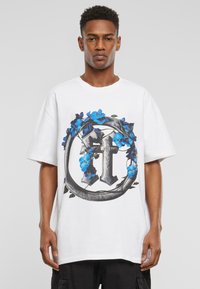 Hvit oversized t-skjorte med et fargerikt grafisk design av et kors omgitt av blå blomster og grønne blader. Korte ermer, rund hals.