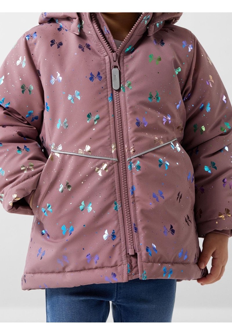 Barn iført en mauve jakke med iriserende sommerfuglmønstre og lynlås foran, hænder delvist i lommerne, parret med blå jeans.