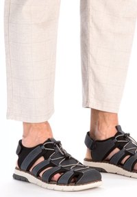 Sandales ouvertes noires et grises avec des lacets élastiques, semelle robuste et coutures contrastées. Portées avec un pantalon à carreaux beige clair, avec un ourlet court.
