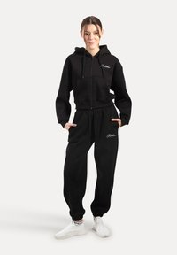 Un sweat à capuche noir avec une fermeture éclair frontale et une poche, associé à un pantalon de jogging noir assorti. Les deux présentent un petit logo. Porté avec des baskets blanches.