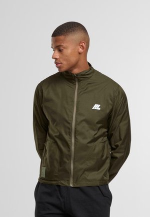 Ung mand ser til venstre iført en olivengrøn zip-up jakke med et hvidt logo på brystet og sorte bukser, hænderne bag ryggen.