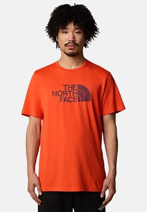 Hombre con pelo rizado que lleva una camiseta naranja con el logo en negro de "The North Face", de pie contra un fondo claro y sencillo.