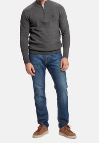 Homme portant un pull à col zippé gris foncé, un jean bleu et des baskets en daim marron, se tenant devant un fond blanc uni.