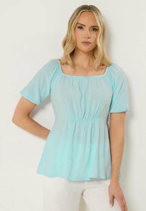 Donna bionda che indossa una blusa azzurro chiaro a maniche corte con vita arricciata e scollo quadrato, in piedi contro una parete bianca.