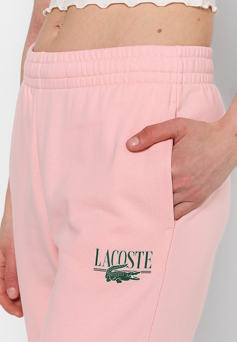 Lichtroze sweatpants met een elastische tailleband, voorzien van een groen Lacoste-logo aan de linkerkant en zijzakken. Zachte stoftextuur.