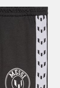 Schwarze Shorts aus weichem Stoff mit einem weißen Streifen, der ein wiederholendes Logo-Muster zeigt, und einem runden Logopatch mit dem Text "MESSI" am Saum.