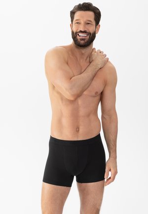 Schwarze Boxer-Shorts aus weichem Stoff, mit nahtlosem Bund und körpernahem Schnitt. Das Modell zeigt einen neutralen Oberkörper und Gesichtsausdruck.